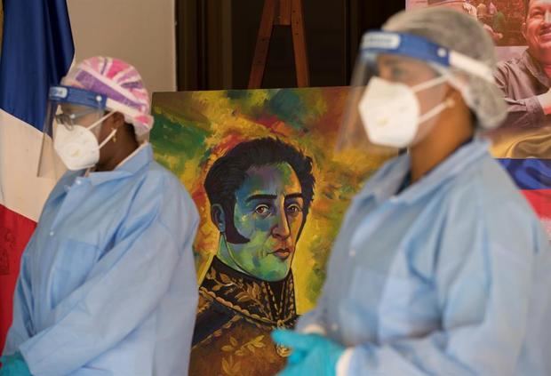 Dos funcionarias de Salud Pública esperan junto a una imagen de Simón Bolívar en la embajada venezolana, donde decenas de ciudadanos de ese país acudieron a realizarse pruebas PCR, este lunes en Santo Domingo.